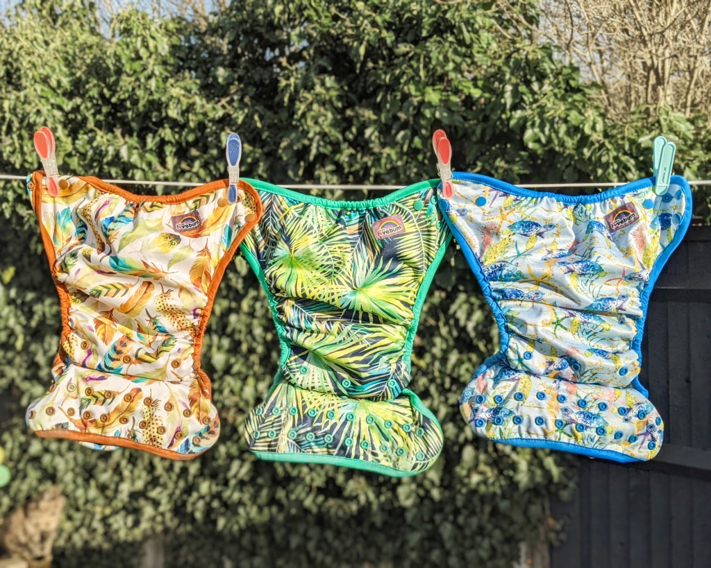 Three colourful reusable nappies hanging on a washing line in a garden in front of a leafy background