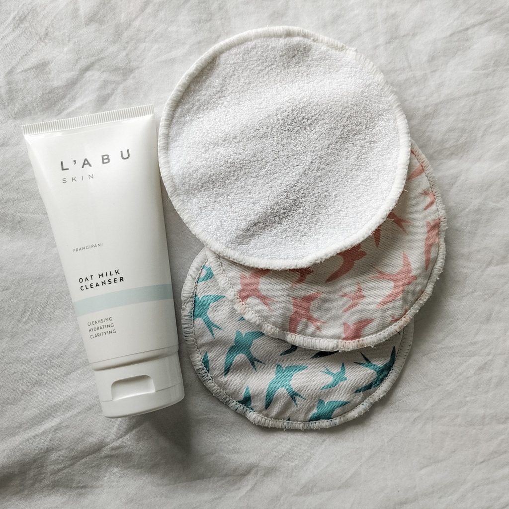 Three round bamboo pads, one face up and two with a patterned reverse side showing with colourful birds on. Next to the wipes lies a bottle of facial cleanser