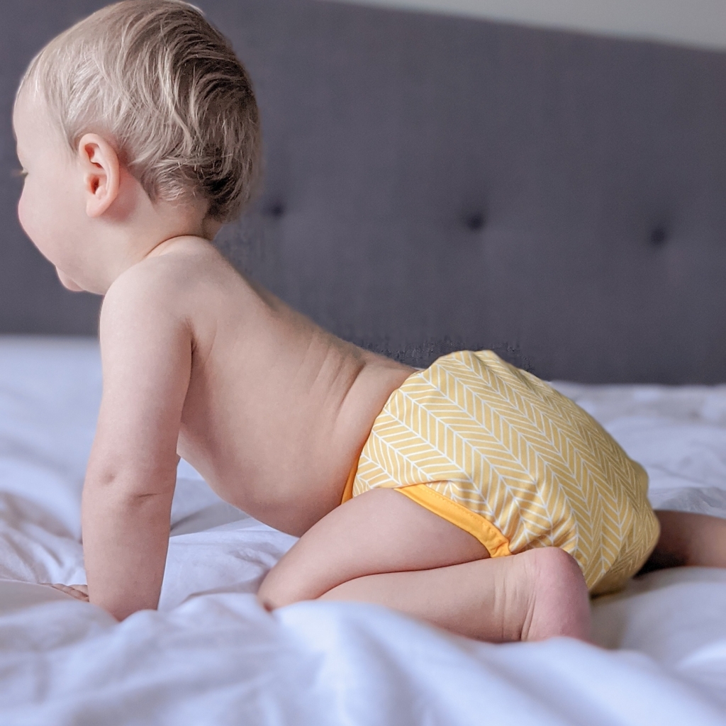 Child is crawling on a bed with white sheets, wearing a yellow cloth nappy in a chevrons pattern.